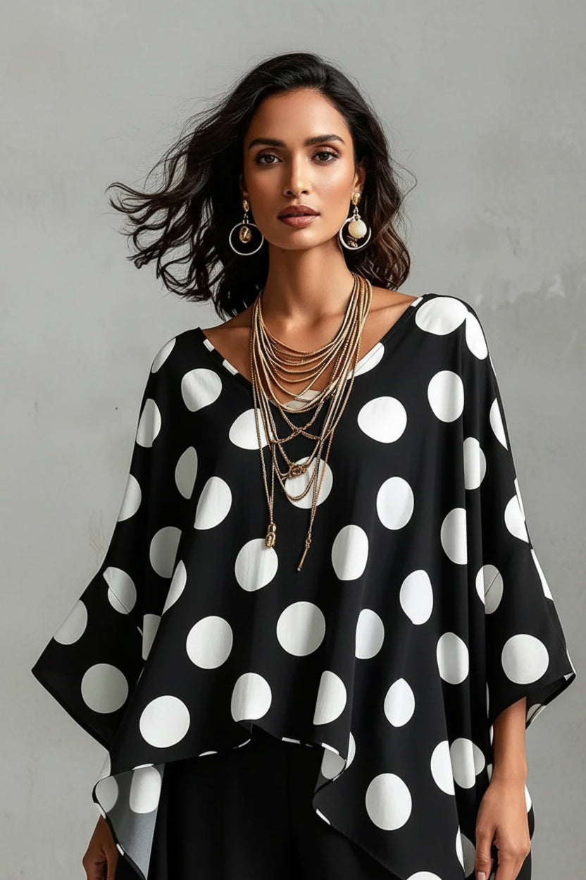 A close-up of a woman wearing a black V-neck top with large white polka dots, accessorized with layered gold necklaces and statement earrings.