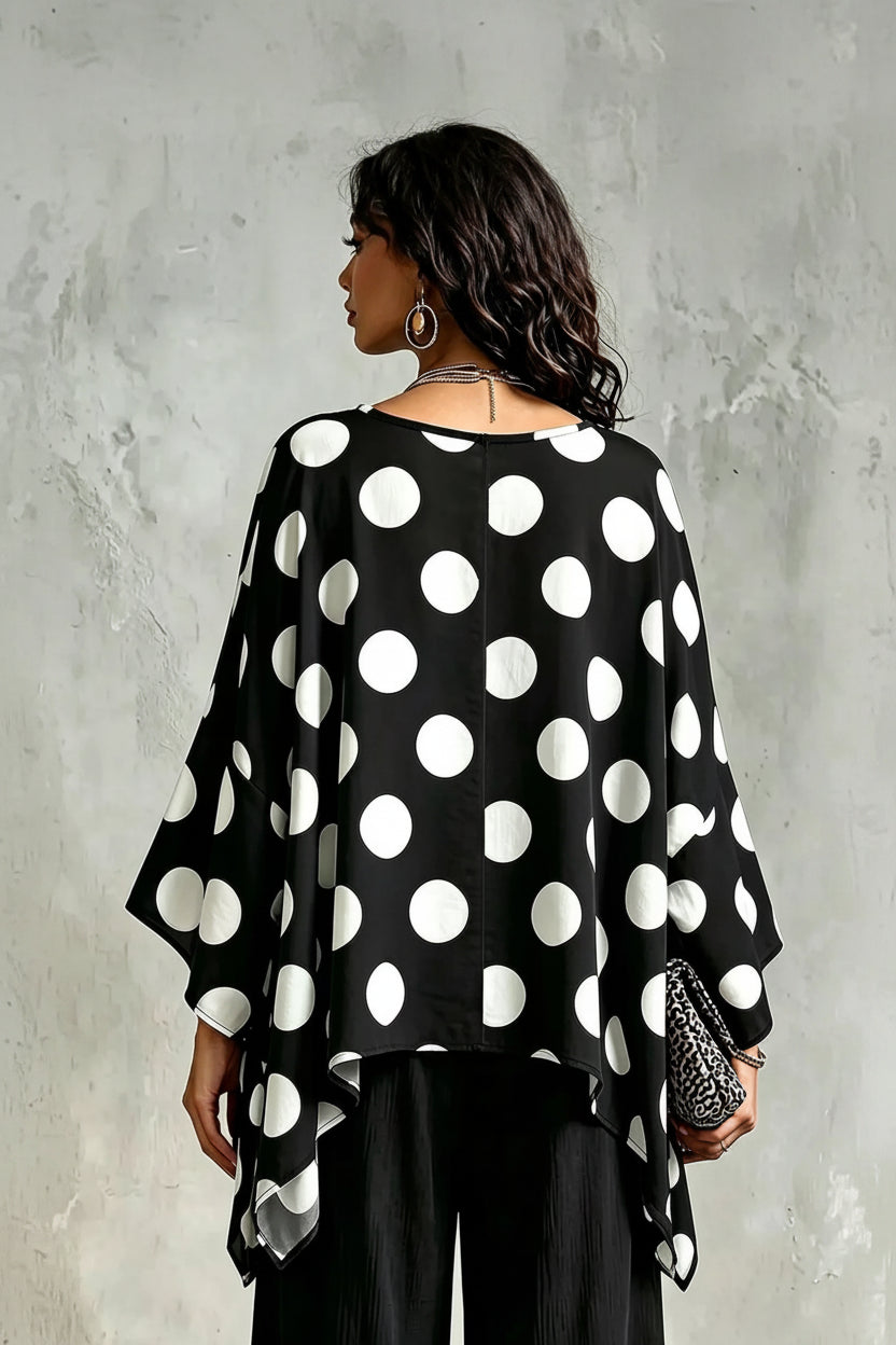A woman shown from the back wearing a loose black top with large white polka dots, paired with black pants, standing against a textured gray wall.