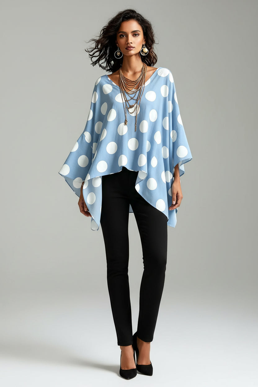A woman wearing a light blue V-neck top with large white polka dots, styled with layered gold necklaces and hoop earrings against a textured gray wall.