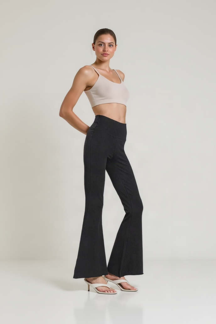 A model stands in a studio wearing black flared pants with a beige crop top and white heeled sandals against a light gray background.