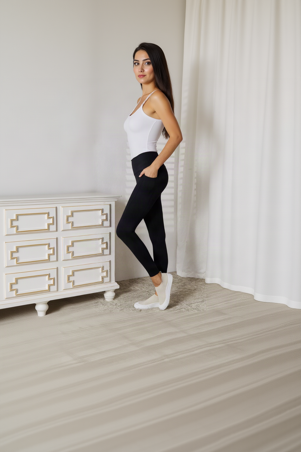 A woman stands in profile wearing a white ribbed tank top and black ribbed leggings with white slip-on shoes, posed in a bright room with a white dresser and curtains.