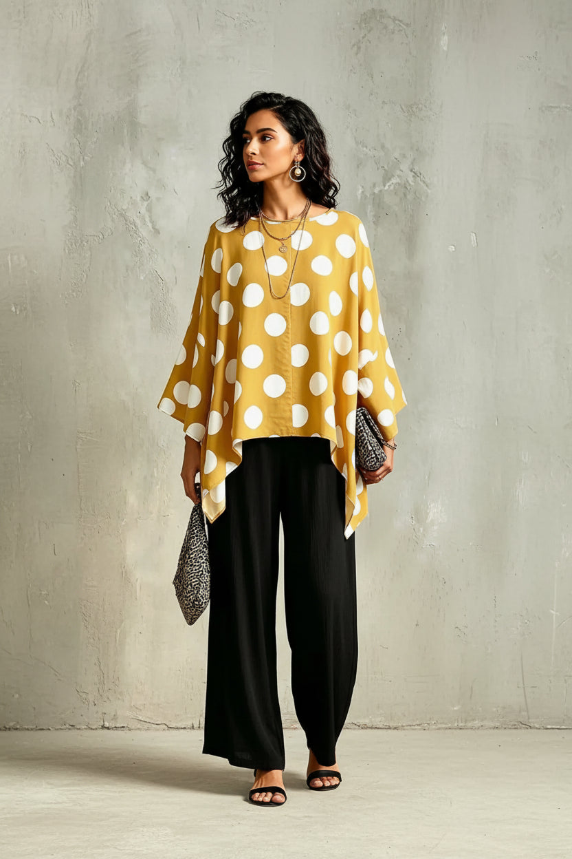 A woman shown from the back wearing a mustard yellow and white polka dot poncho top with wide draped sleeves, paired with black pants, against a textured gray wall.
