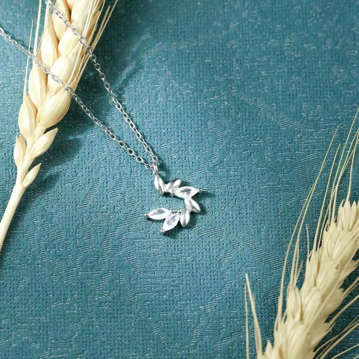 Silver necklace with leaf pendant on a textured teal surface with wheat stalks.