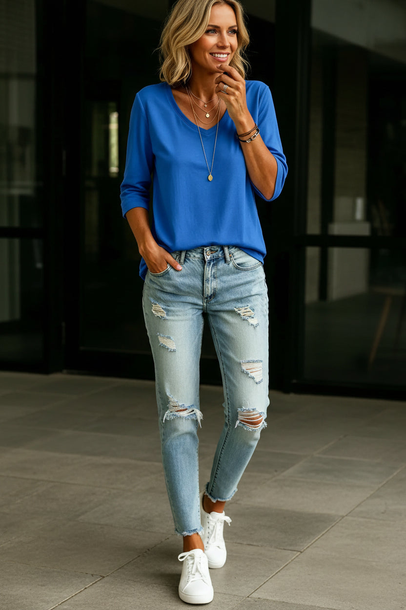 A woman wearing a bright blue three-quarter sleeve V-neck top and light-wash distressed denim jeans, accessorized with layered gold necklaces and bracelets.