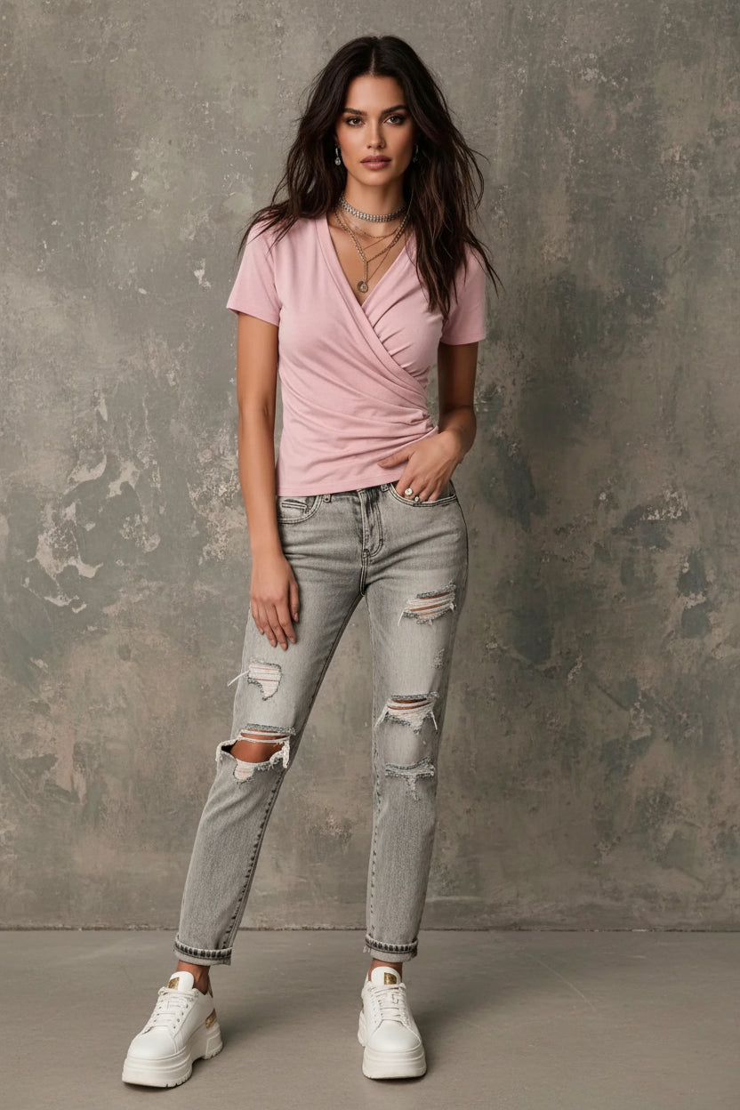 A woman wearing a light pink short-sleeve wrap-style V-neck t-shirt, styled with layered silver necklaces and light-wash jeans against a textured gray background.