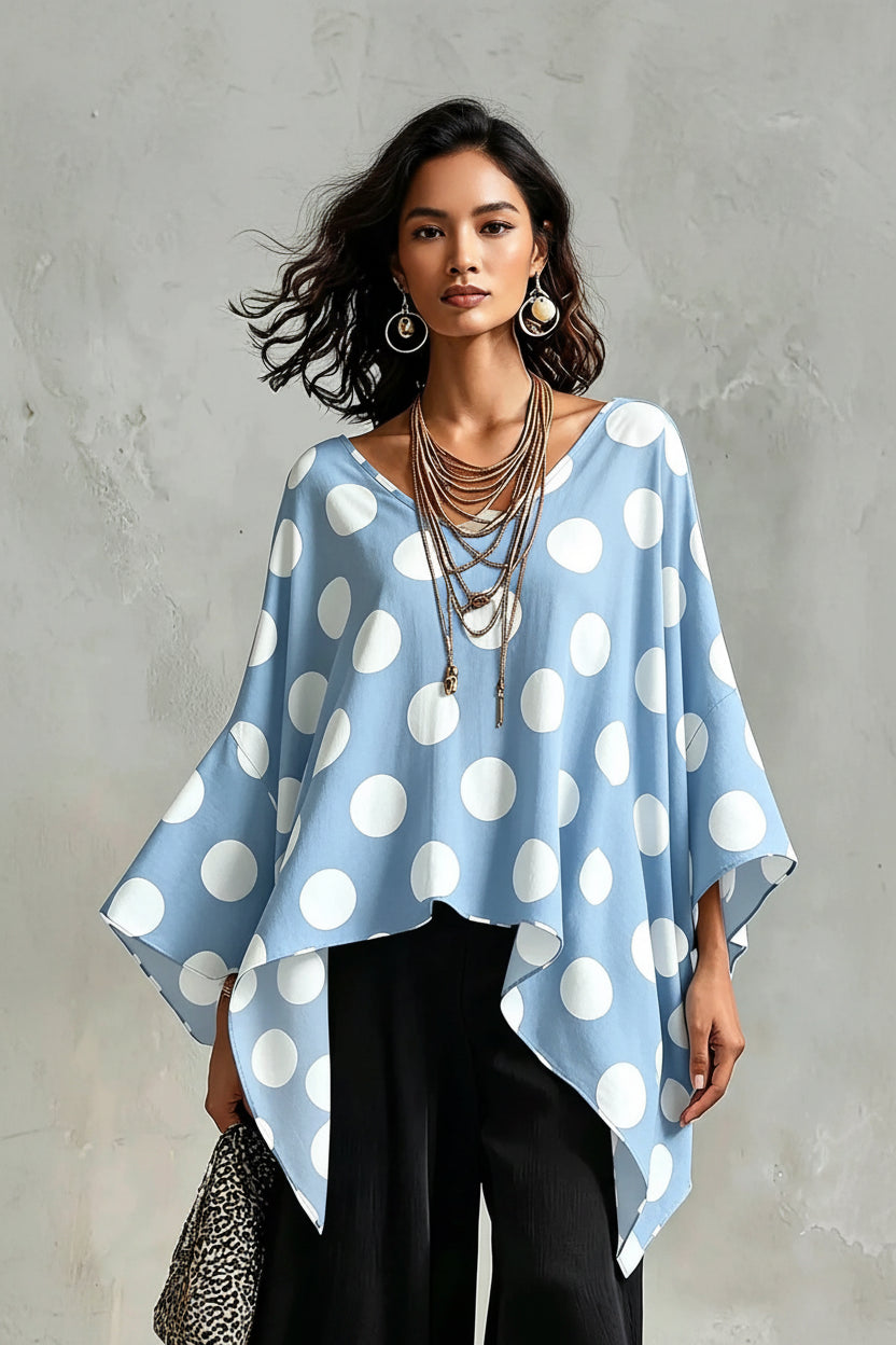A woman wearing a light blue and white polka dot poncho top with wide bell sleeves, paired with black pants and layered gold necklaces, standing against a textured gray wall.