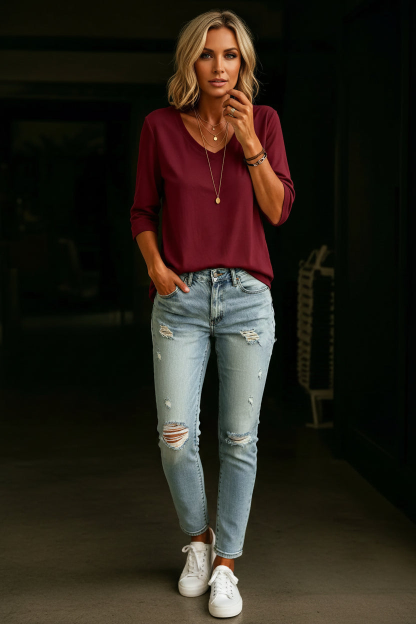 A woman wearing a burgundy long-sleeve V-neck top and light-wash distressed denim jeans, accessorized with layered gold necklaces and bracelets.