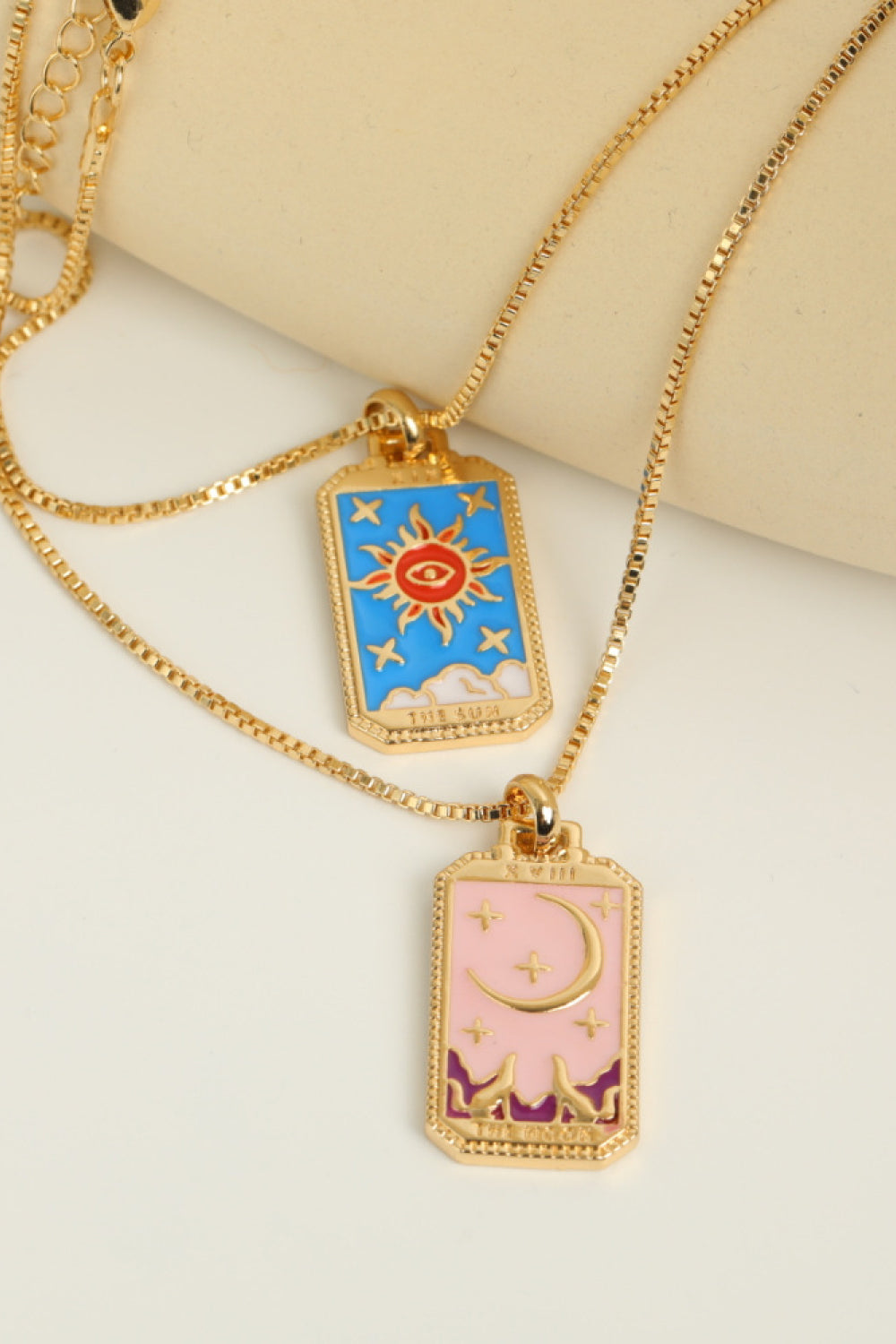 Two gold necklaces with colorful pendant designs on a beige background