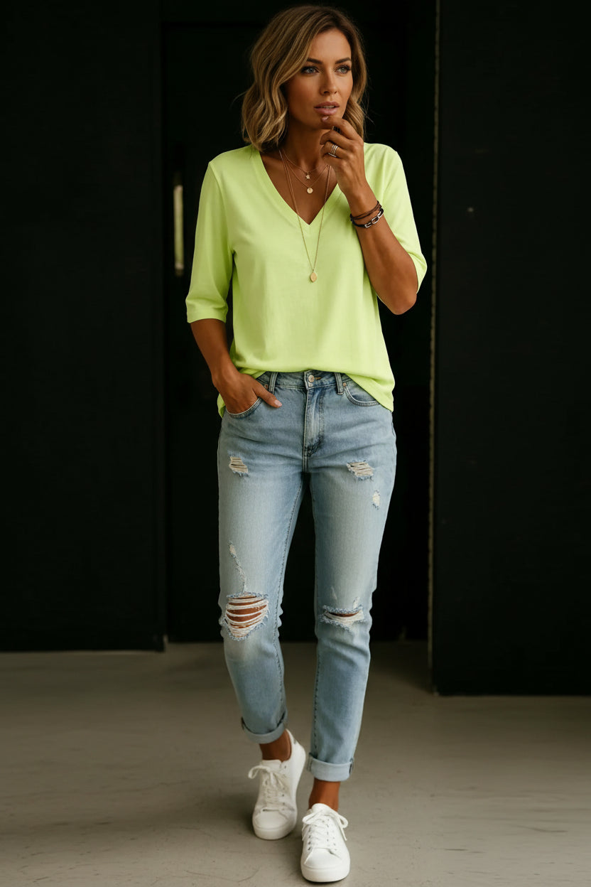 A woman wearing a light lime green V-neck short-sleeve top and light-wash distressed blue jeans, accessorized with layered gold necklaces and bracelets.