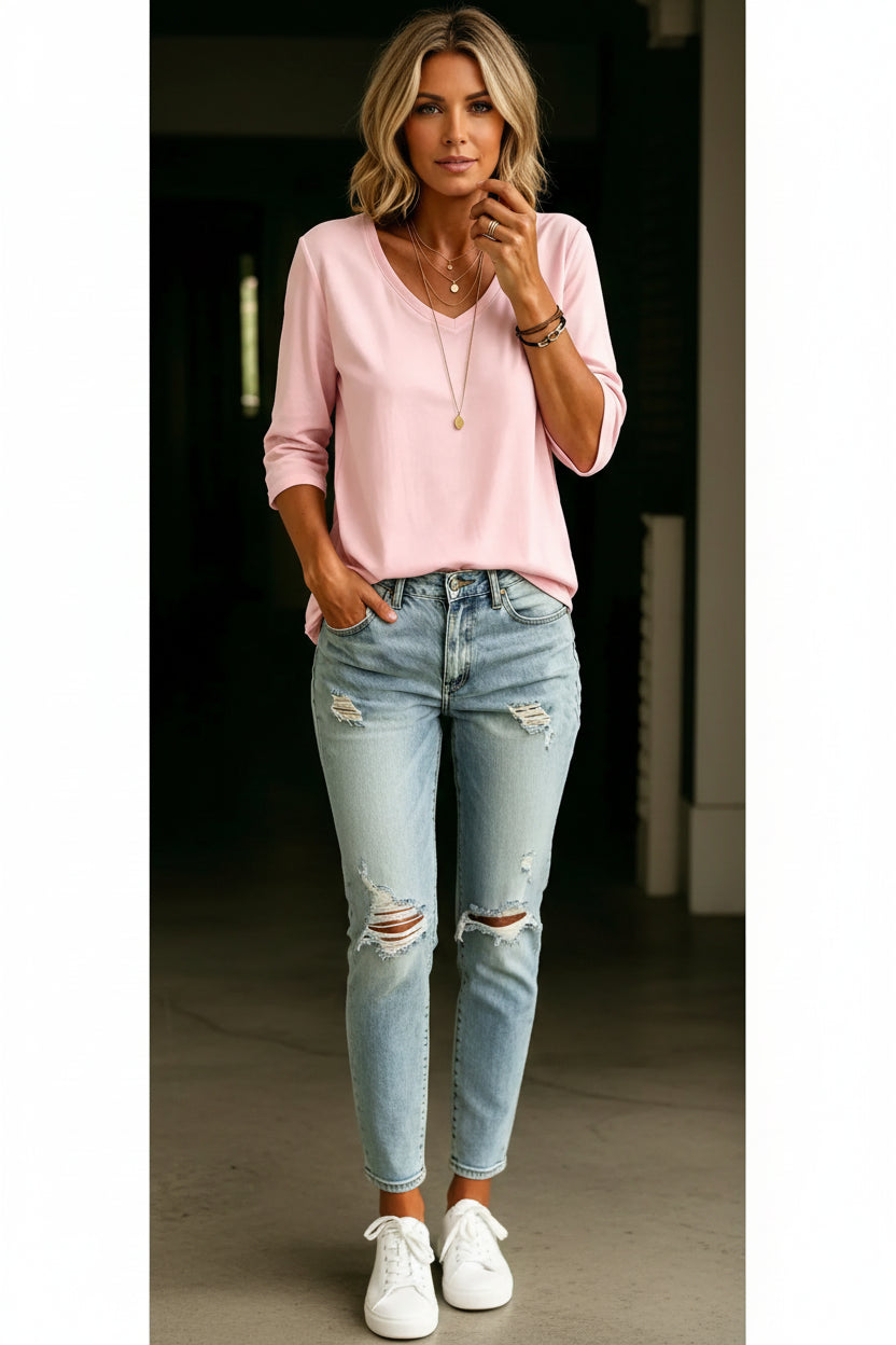 A woman wearing a light pink long-sleeve V-neck top tucked into light-wash distressed denim jeans, accessorized with layered gold necklaces and bracelets.