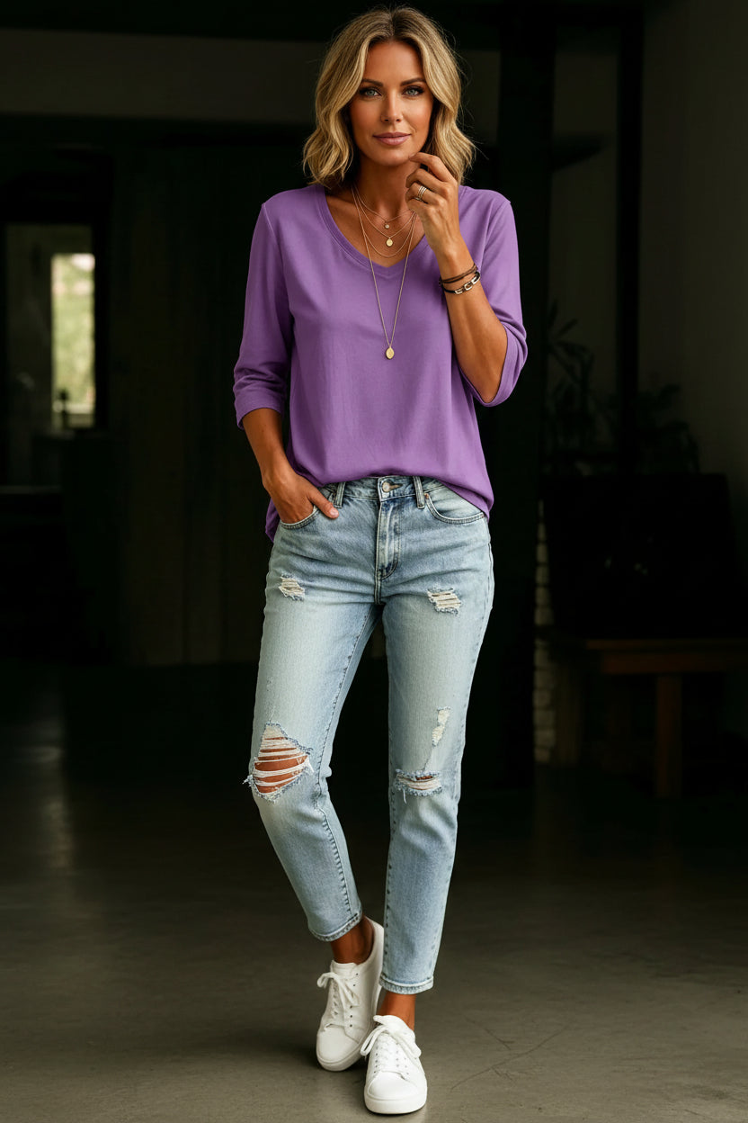 A woman wearing a purple three-quarter sleeve V-neck top and light-wash distressed denim jeans, accessorized with layered gold necklaces and bracelets.