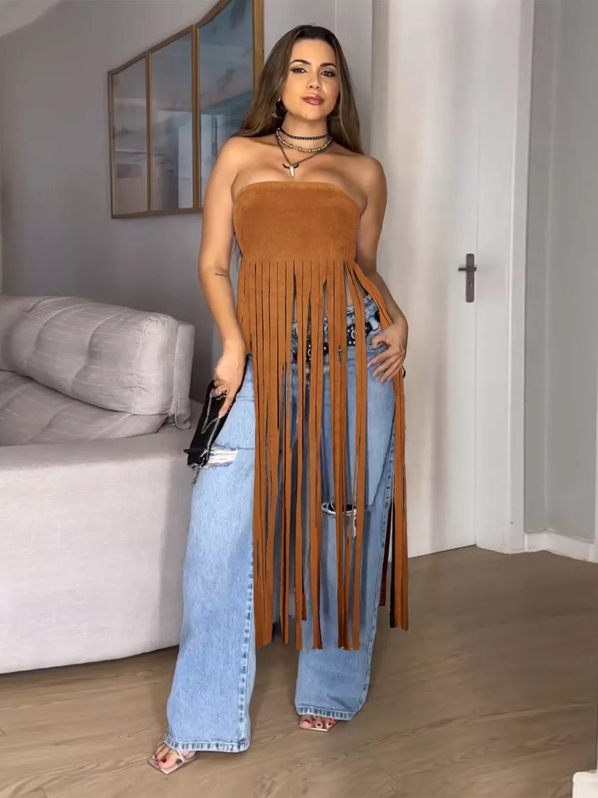 Woman wearing a brown fringe top and blue jeans in a room with a couch and mirror.