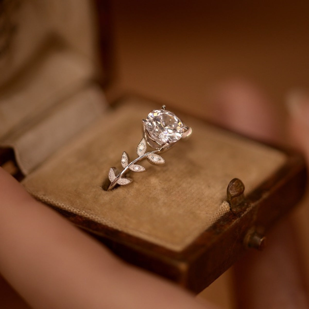 Diamond ring with leaf design on a brown velvet cushion in a wooden box.