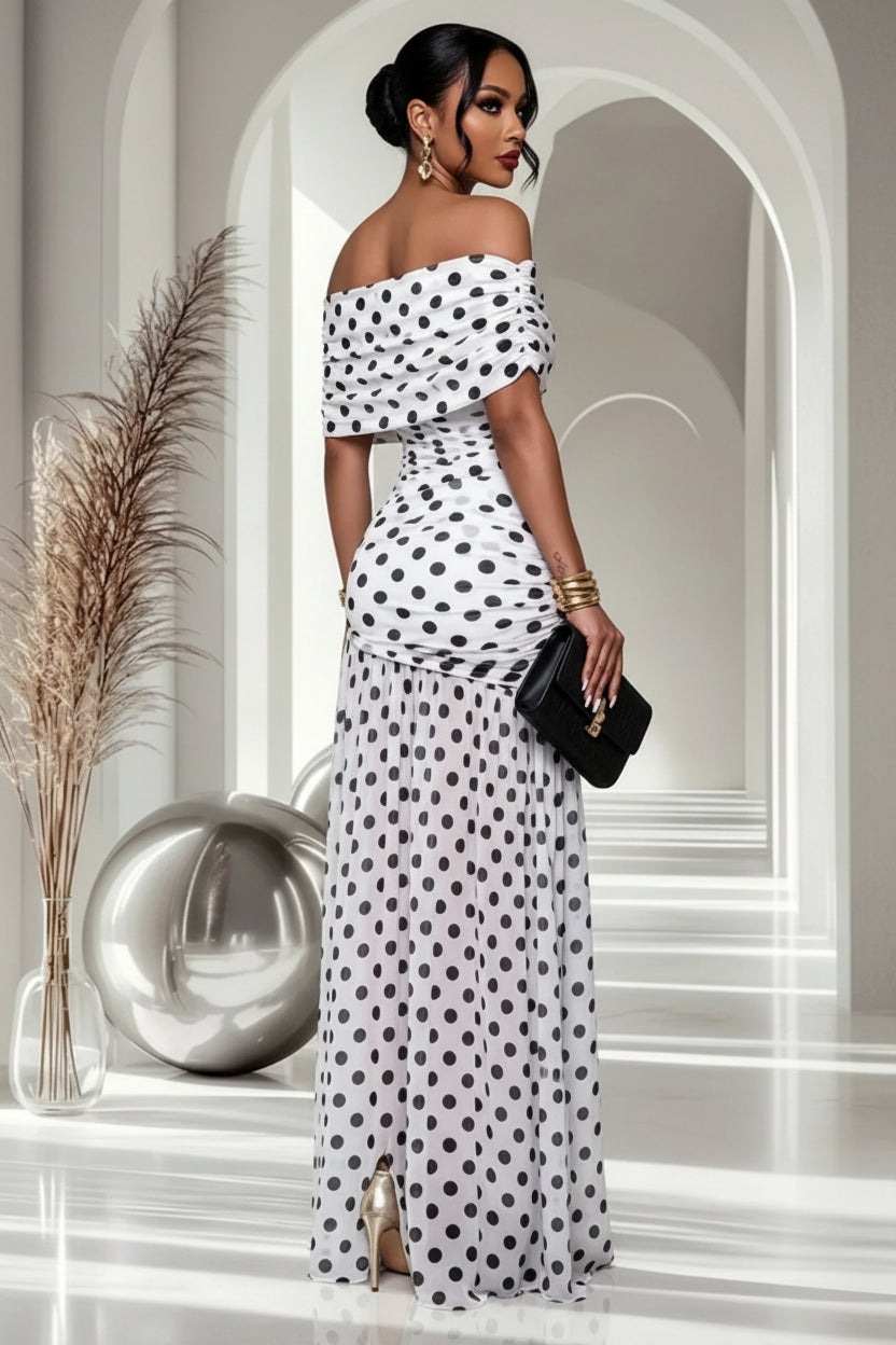A woman modeling an off-the-shoulder white maxi dress with black polka dots, shown from the back in a bright, modern hallway with decorative pampas grass and a metallic sphere.