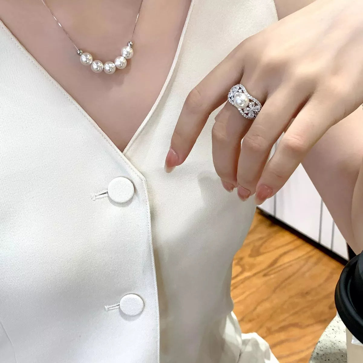 Close-up of a person wearing a pearl necklace and ring, with a white blouse and wooden floor background.