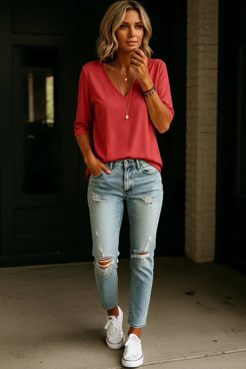 A woman wearing a coral red V-neck short-sleeve top and light-wash distressed denim jeans, accessorized with layered gold necklaces and bracelets.