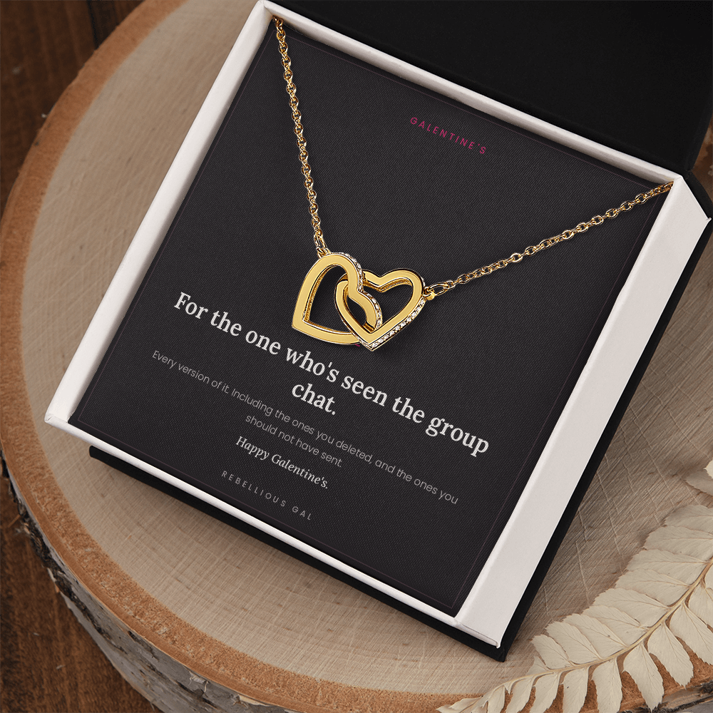 Gold interlocking heart necklace in a white box on a wood slab beside dried fern