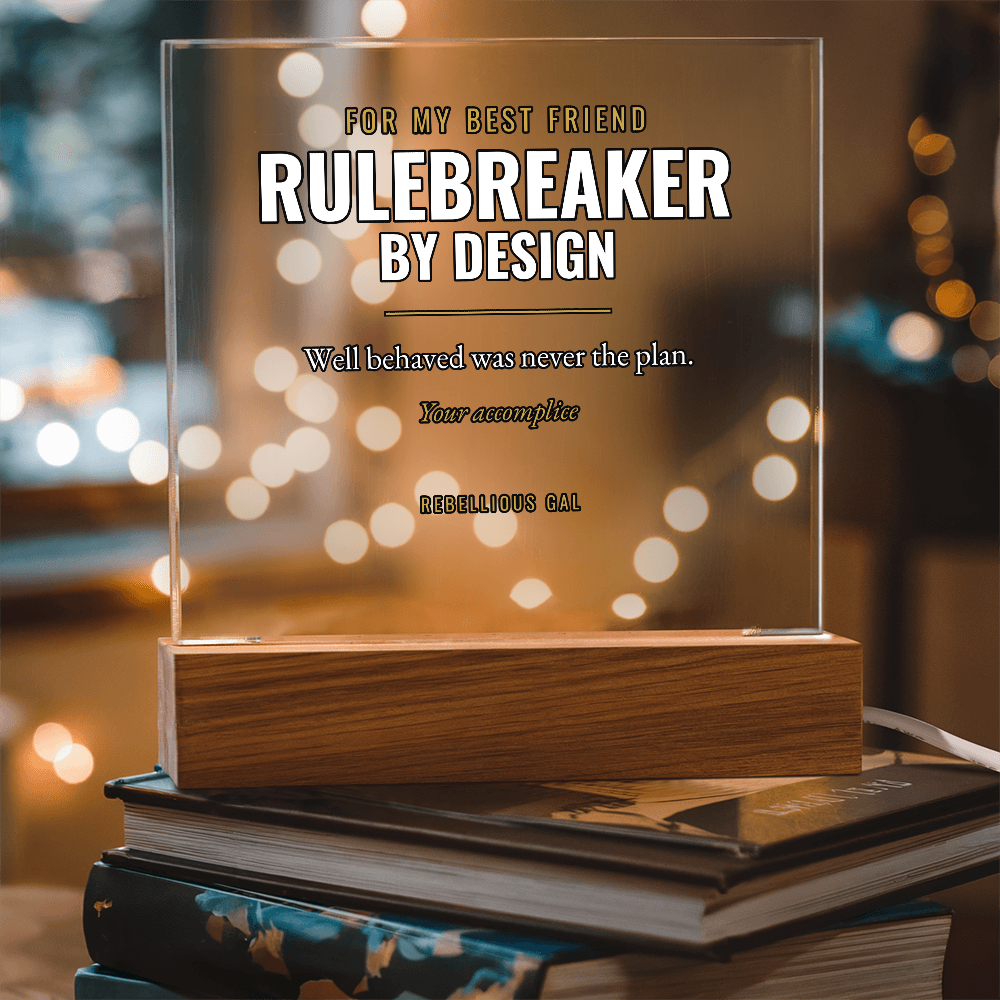 Clear acrylic Rulebreaker plaque on a wooden lamp base atop stacked books, bokeh glow