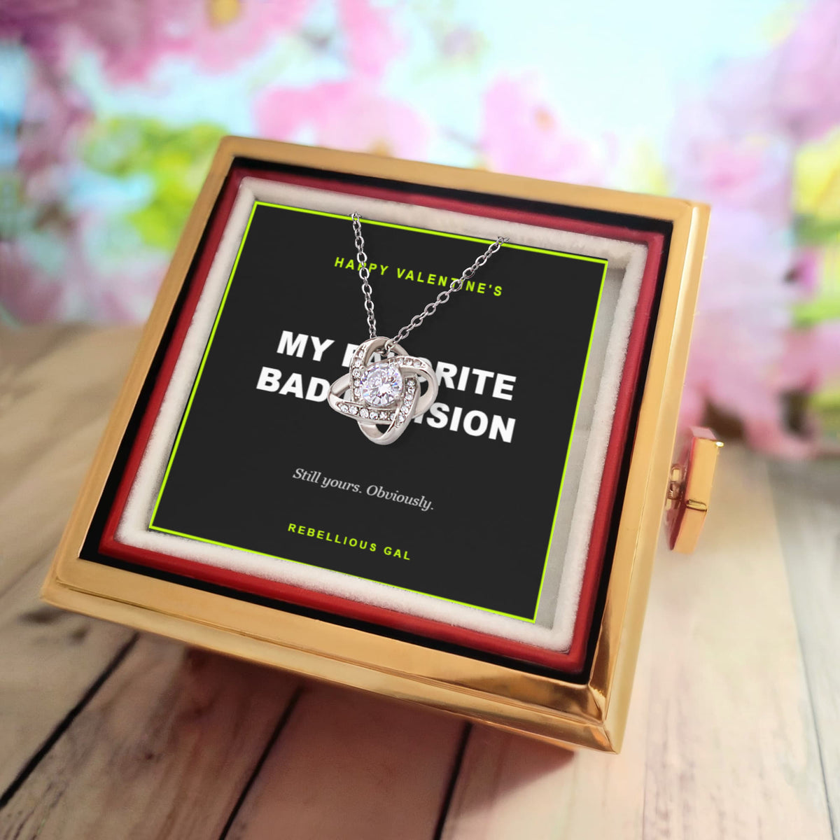 Gift box on pale wood boards, blurred pink blossom behind, silver knot pendant on its card