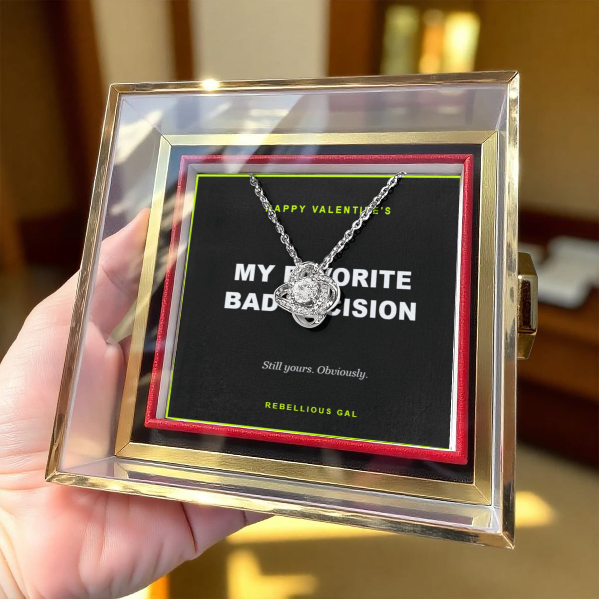Hand holding the gold-trimmed box up indoors, silver love knot pendant on a black card