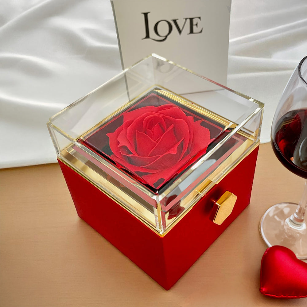 Closed red box with a red rose under the clear lid, beside a glass of wine