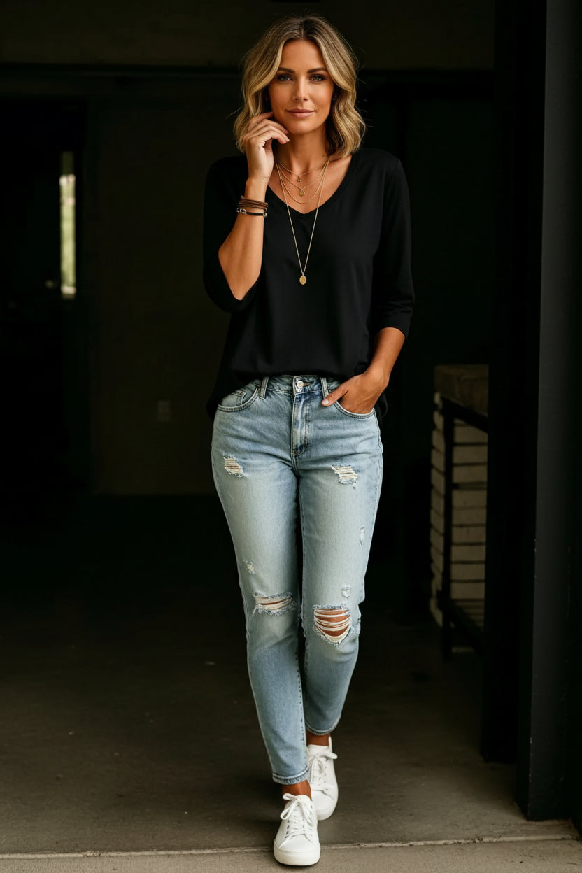 A woman wearing a black three-quarter sleeve V-neck top and light-wash distressed denim jeans, accessorized with layered gold necklaces and bracelets.