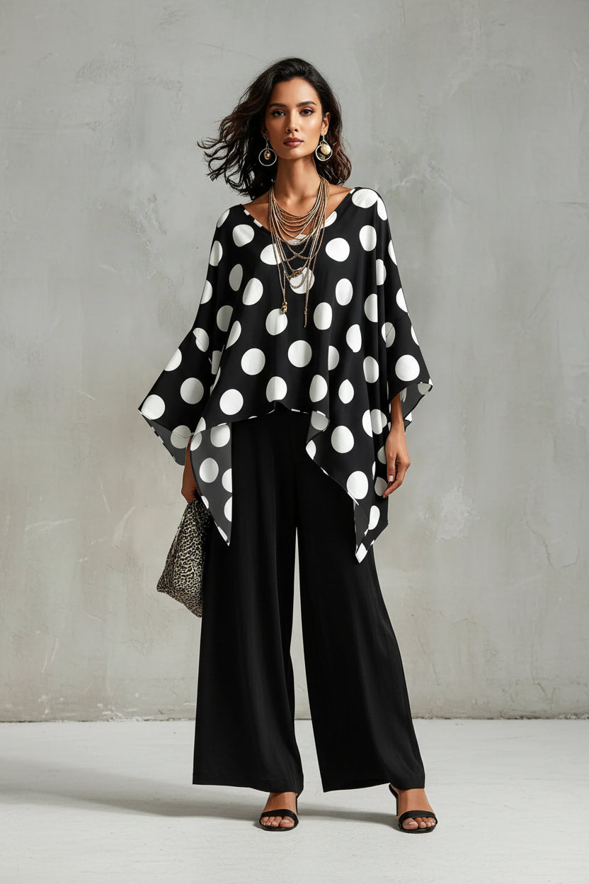 A woman wearing a black V-neck poncho-style top with large white polka dots, layered with multiple long necklaces, against a textured gray background.