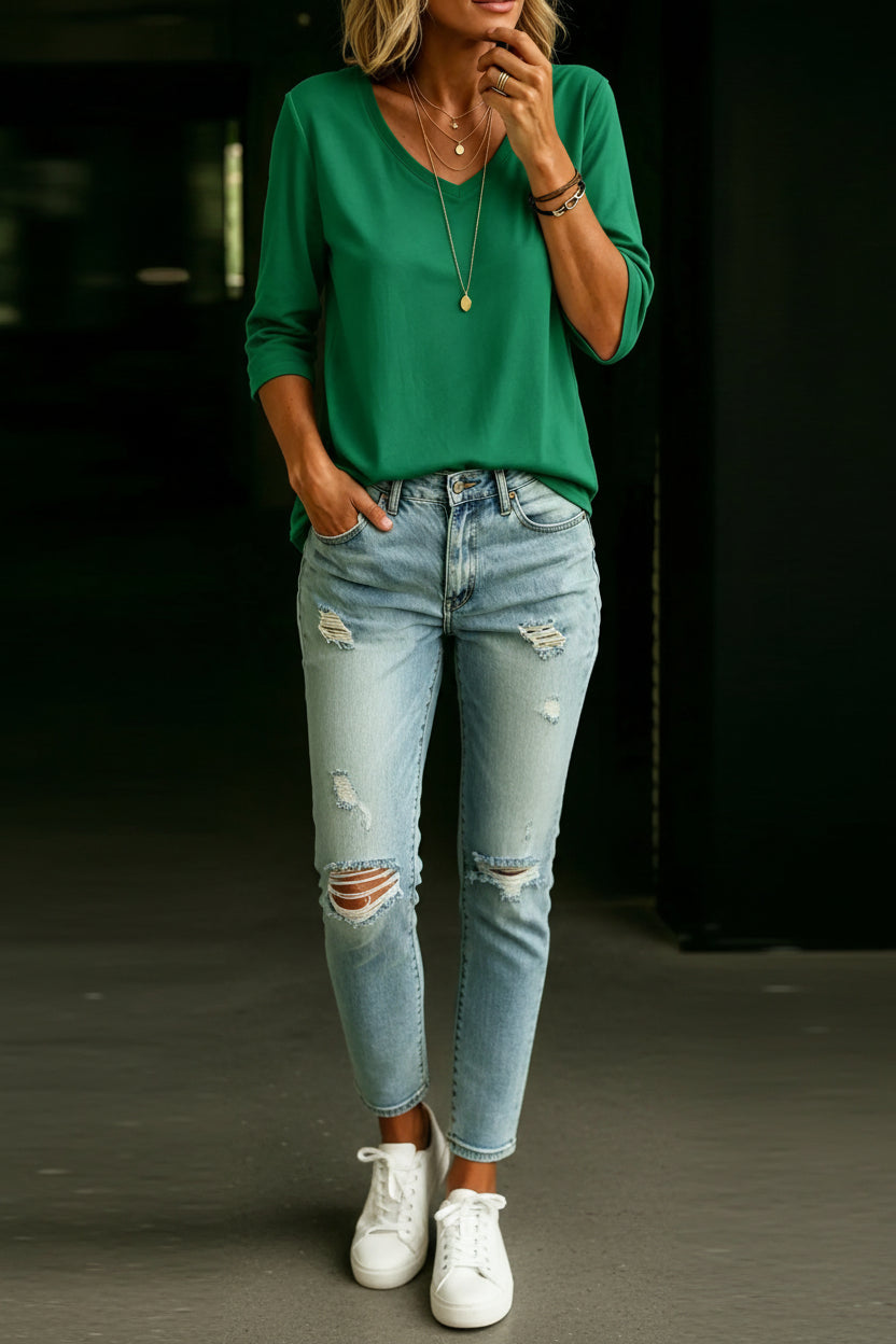 A woman wearing a vibrant green long-sleeve V-neck top and light-wash distressed denim jeans, accessorized with layered gold necklaces and bracelets.