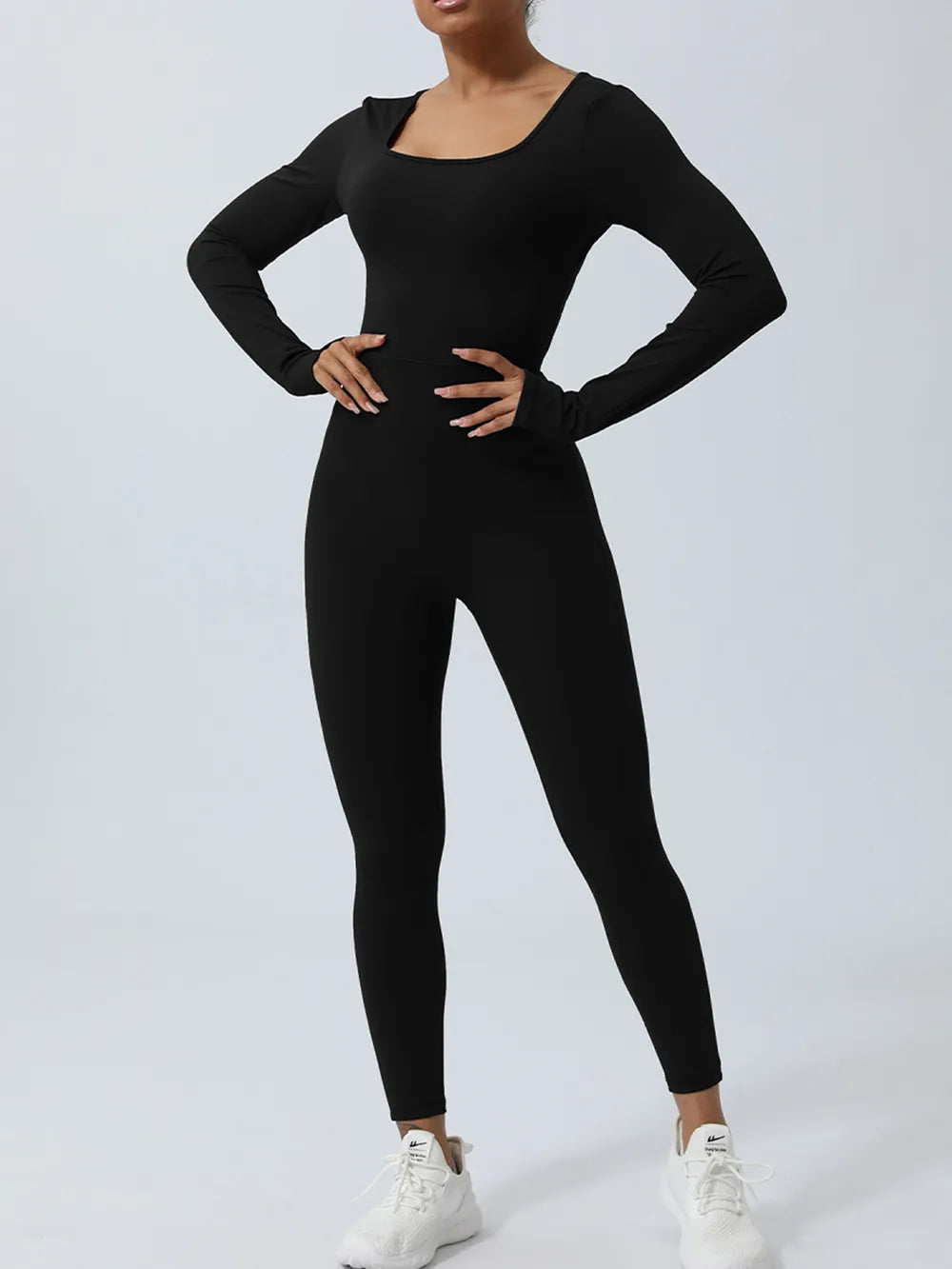 Black long sleeve jumpsuit, scooped neckline, both hands on the hips above white sneakers