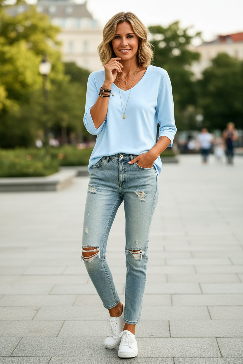 A woman wearing a light blue V-neck long-sleeve top tucked into light-wash distressed denim jeans, accessorized with layered gold necklaces and bracelets.
