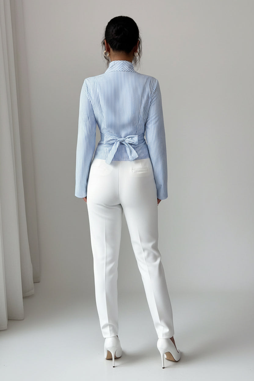 A woman shown from the back wearing a light blue and white vertically striped long-sleeve blouse with a tie detail at the waist, paired with white pants.