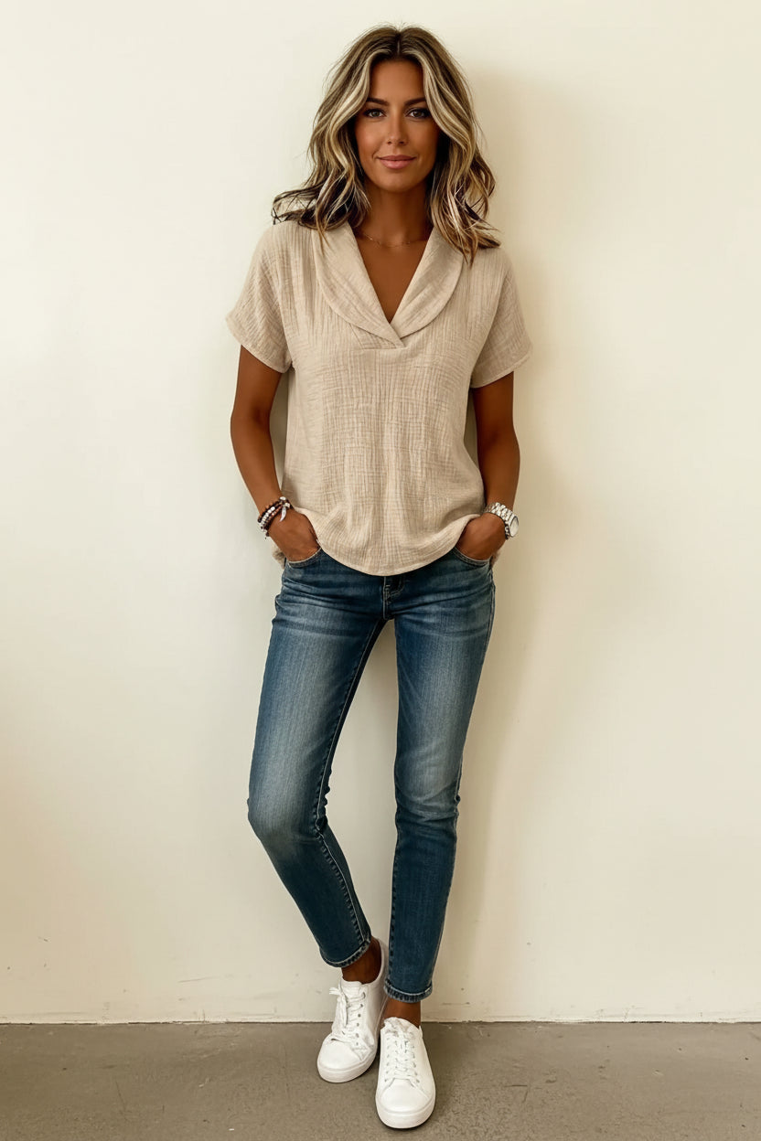A woman wearing a light beige short-sleeve top with a draped cowl neckline, paired with blue jeans, standing against a plain light beige wall.