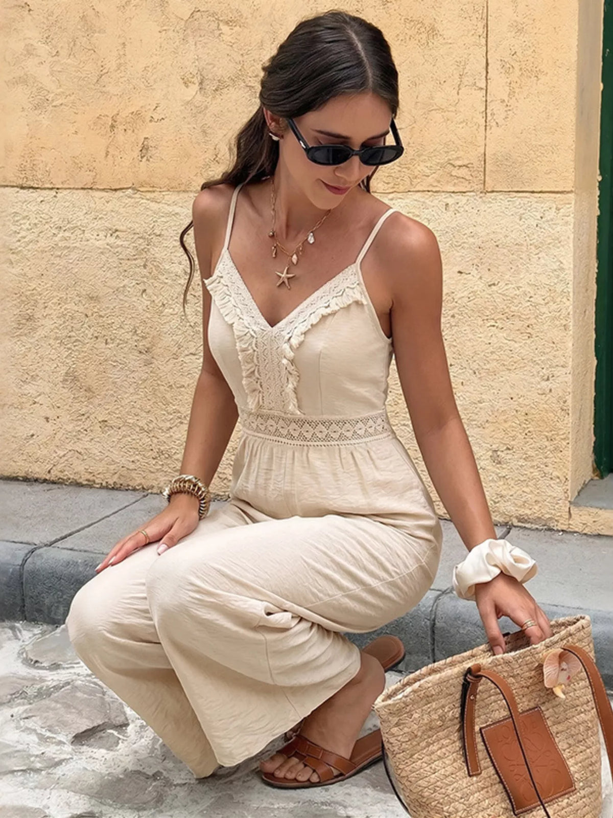 Woman in a beige dress with sunglasses and a straw bag against a stone wall.