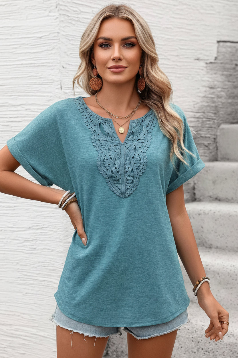 Woman wearing a teal short-sleeve top with lace detail against a textured white wall.