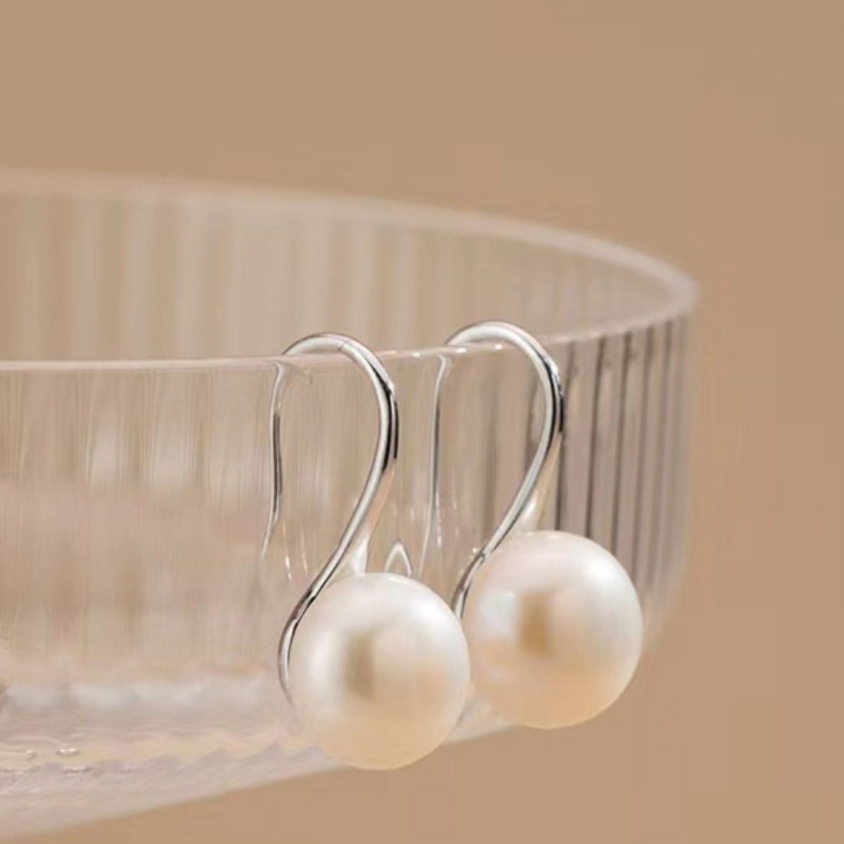 Pearl earrings hanging from a clear glass bowl on a beige background