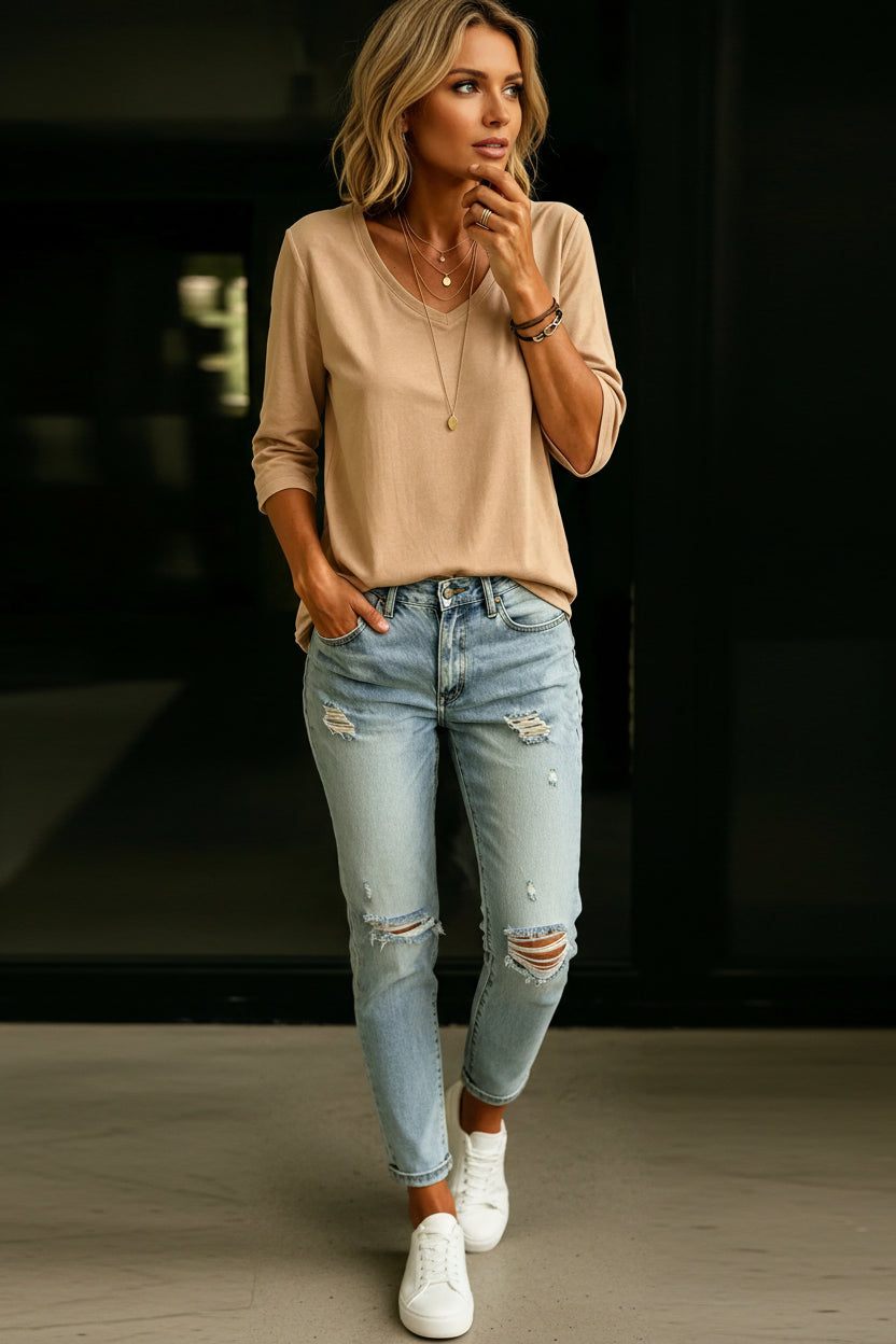 A woman wearing a beige long-sleeve V-neck top and light blue distressed denim jeans, accessorized with layered gold necklaces and bracelets.