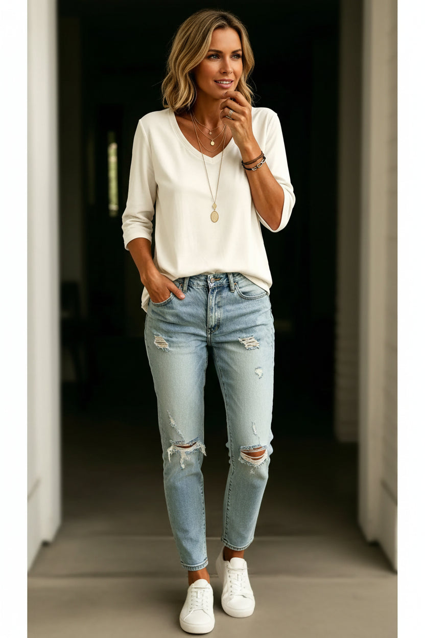 A woman wearing a cream-colored, long-sleeve V-neck top tucked into light-wash distressed denim jeans, accessorized with layered gold necklaces and bracelets.