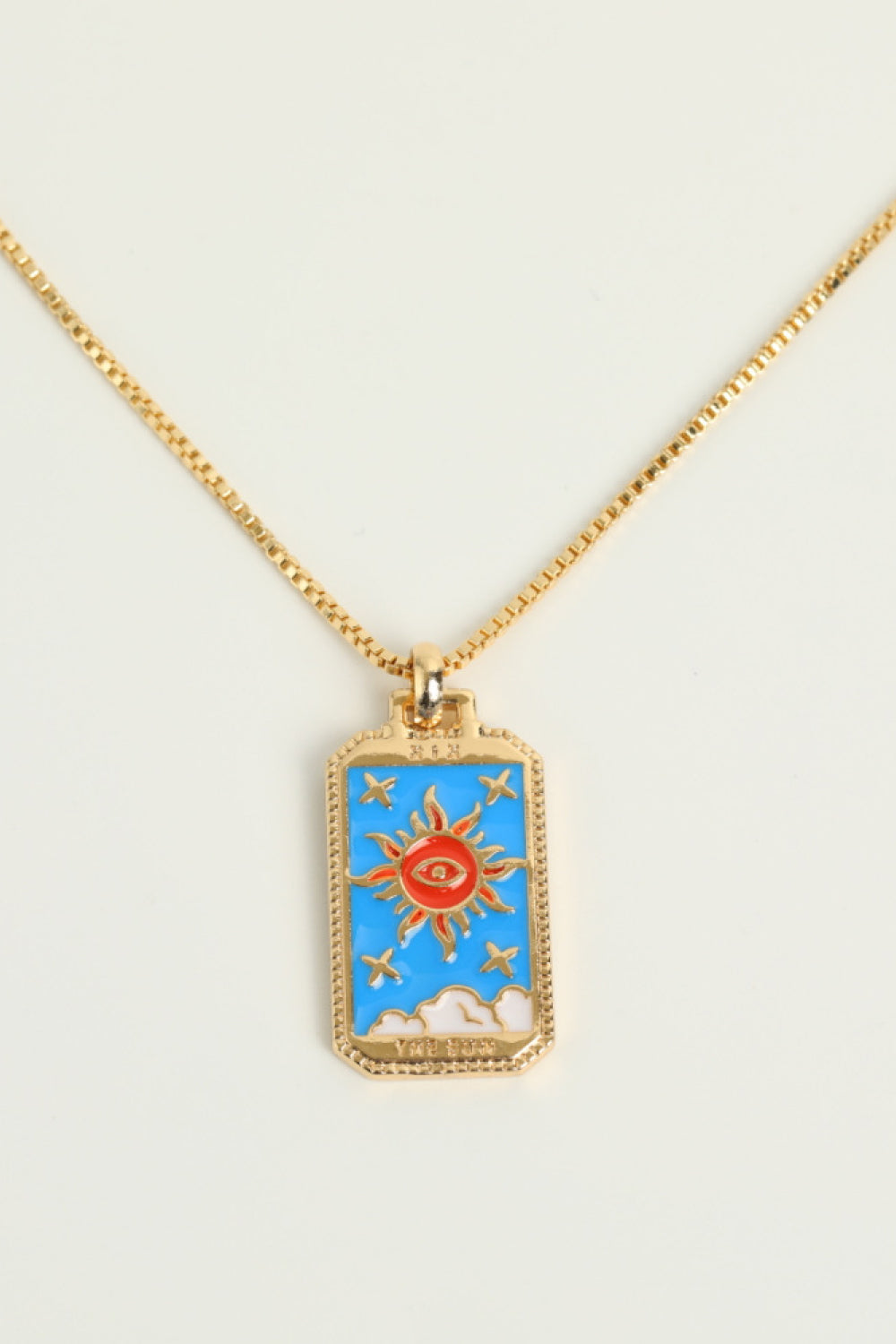 Gold necklace with a rectangular pendant featuring a sun and eye design on a white background