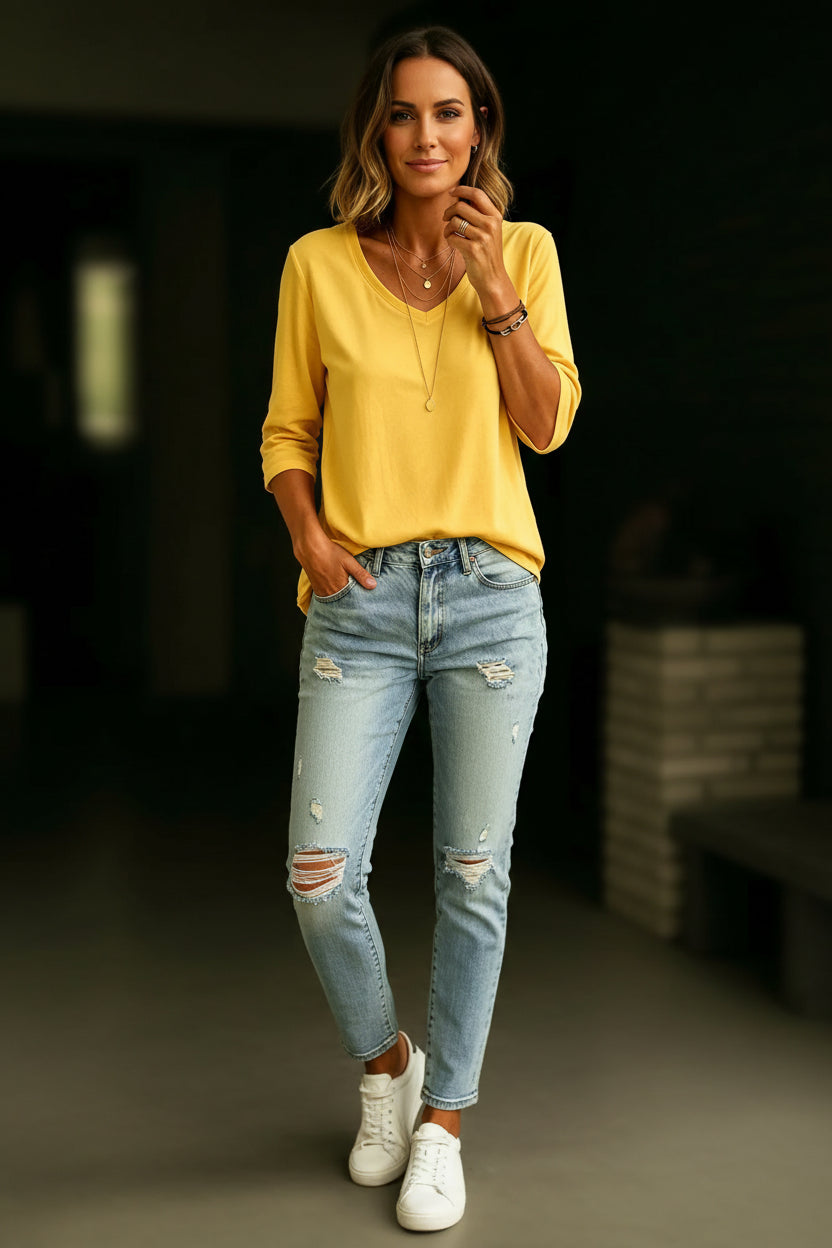 A woman wearing a yellow V-neck long-sleeve top and light blue distressed jeans, accessorized with layered gold necklaces and bracelets.